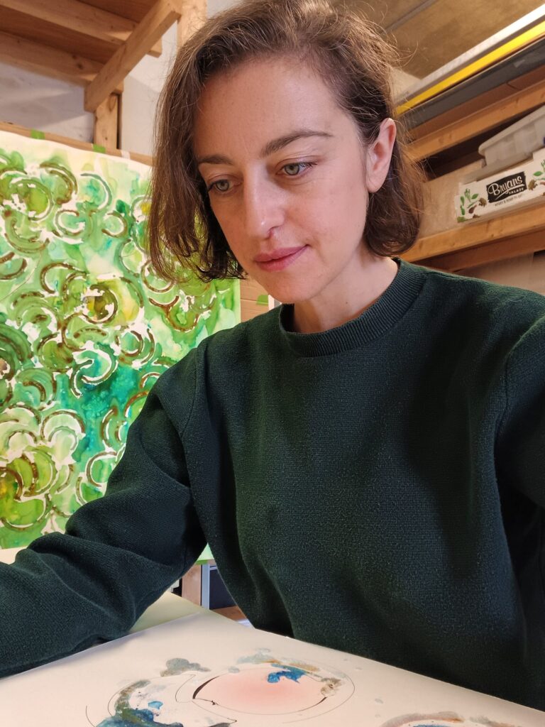 profile picture of a woman in a green jumper with a painting behind her and in front of her on the table